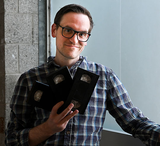 headshot of man with glasses, holding VHS tapes
