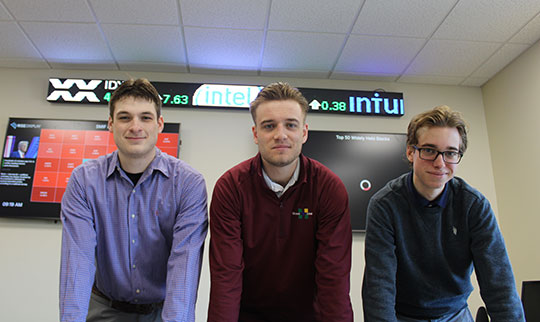 three young men standing in a row, with stock trade ticker behind them