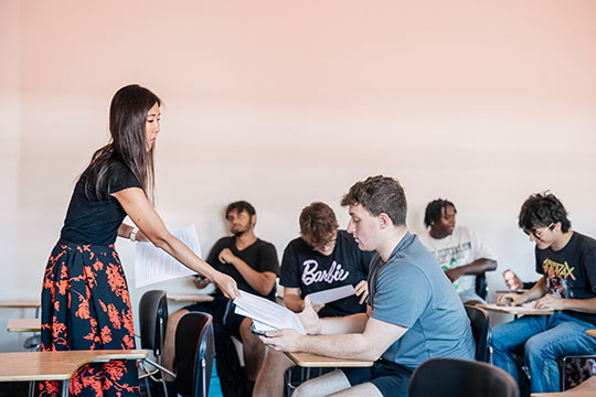 woman in dress handing out papers to students in a classroom