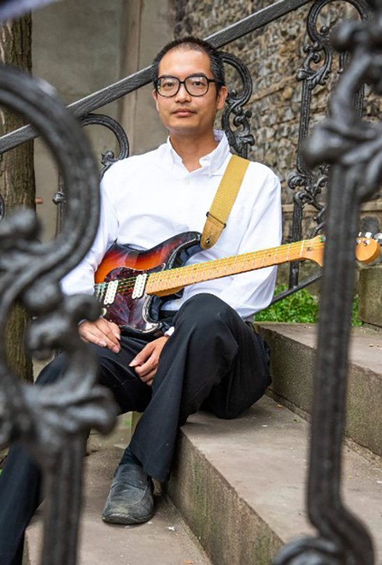 Ken Luk sitting on steps holding an electric guitar.