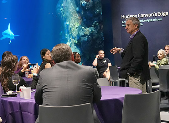 man in sport coat talking to a crowd, standing in front of aquarium