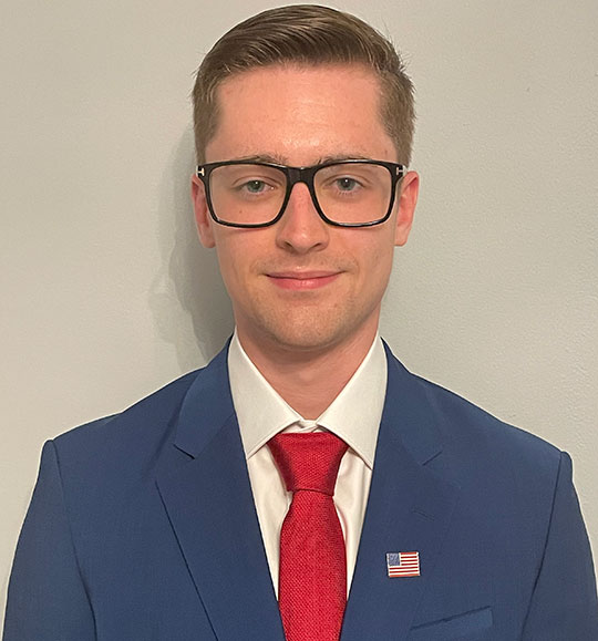 headshot of young man with coat and tie, glasses
