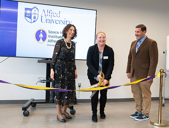 three people, two women and a man, one woman using giant scissors to cut a ribbon