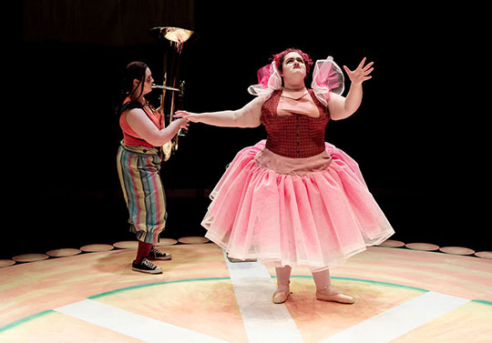 woman on stage acting, someone playing tuba behind them