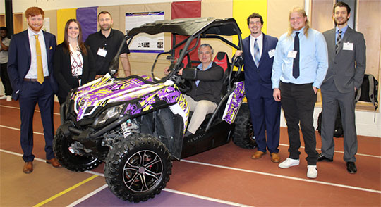 A group of seven people, dressed in business attire, pose around a sporty, purple off-road vehicle indoors. The mood is celebratory and professional.