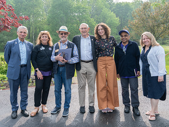 A group of seven people stand smiling outside on a paved path, surrounded by lush greenery and trees. The mood is cheerful and relaxed.
