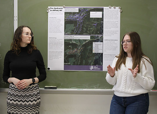 Two individuals stand in front of a green chalkboard presenting a poster titled "Fire Hydrants in Alfred, NY." The poster includes maps and text. One person is speaking, gesturing with hands, while the other looks on. They appear focused and engaged.