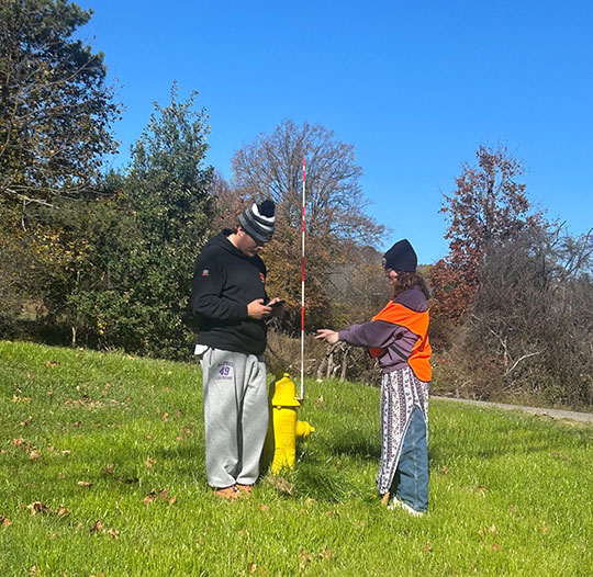 Two people stand near a yellow fire hydrant in a grassy area. One points upward, wearing an orange vest, while the other looks at their phone. It's a sunny day.