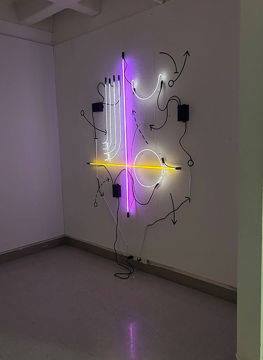 Neon glass artwork titled ‘Game Plan’ by Alfred University student Amelia Booth, featuring glowing purple, yellow, and white lines forming sports play diagrams on a wall.