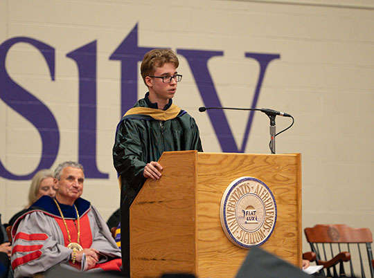 A young man in a graduation gown speaks at a podium with a university insignia. Behind him, faculty members in academic regalia sit, listening attentively.