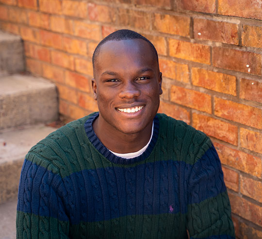 young man in sweater smiling, brick wall and steps behind him