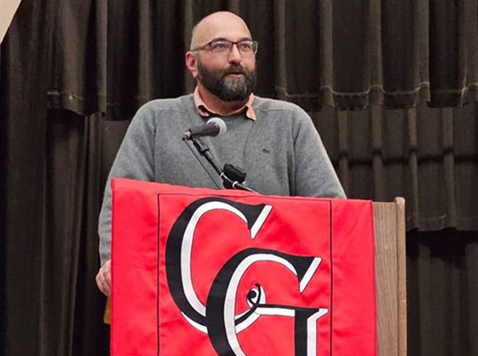 A man stands at a podium draped with a red cloth featuring the letters "CG." He is speaking into a microphone, with a serious expression. Dark curtains in the background.