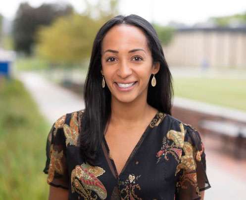 Smiling woman with long dark hair and dangling earrings, wearing a black floral blouse. She's outdoors with a blurred green and brown background.