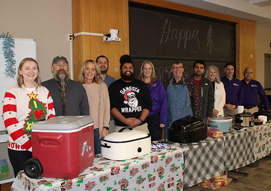 A diverse group of eleven people stands smiling behind a table with festive tablecloths, coolers, and food trays. A chalkboard behind them reads "Happy." The atmosphere is cheerful and celebratory.