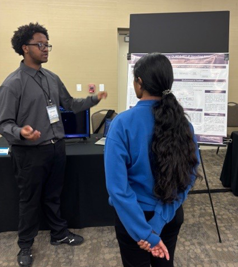 Man in a gray shirt presents a scientific poster to a woman in a blue sweater at a conference. The setting is professional and focused.