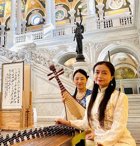 Two women in traditional attire play string instruments in a grand, ornate hall with marble stairs and murals. The scene feels elegant and cultural.