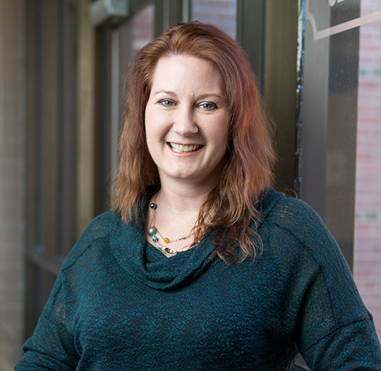 Alfred University psychology professor standing by a window, smiling, wearing a teal sweater and necklace.