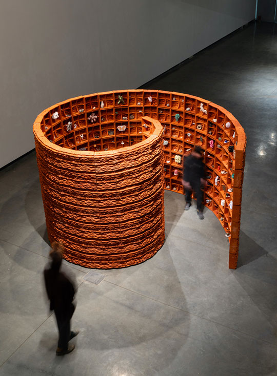 Overhead view of a spiral installation made of textured orange shelves filled with small objects. Two blurred people walk around it on a concrete floor.