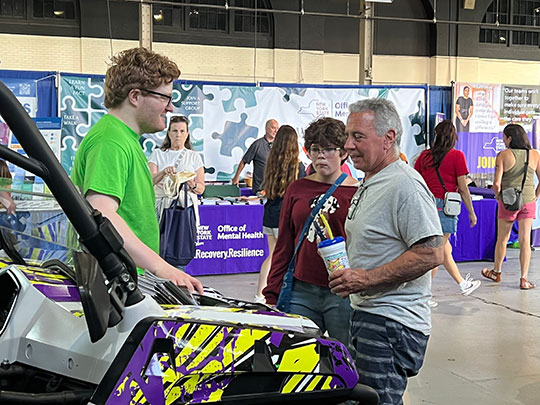 A man in a green shirt converses with a person holding a drink by a colorful vehicle at an indoor event. Booths with mental health banners are in the background.