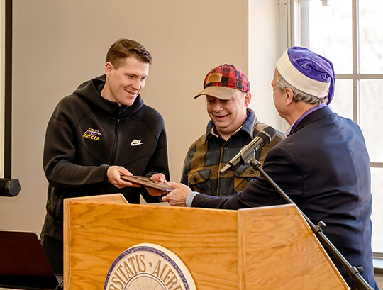 three men at a podium, one wearing a santa claus hat and presenting plaques to the other two