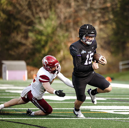 Alfred University football player in a black uniform running with the ball while an opposing player in a white and red uniform attempts to tackle him.