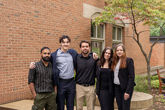 A group of five students dressed in business casual attire stand together outside an Alfred University building, smiling for a photo.