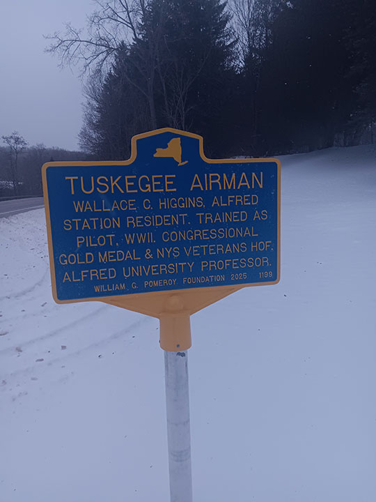 Blue and yellow historical marker in a snowy landscape honors Tuskegee Airman Wallace C. Higgins, highlighting his achievements and affiliations.