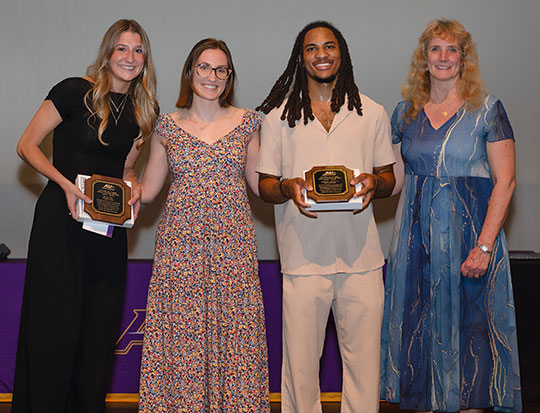 four people standing in a row, smiling, two holding awards
