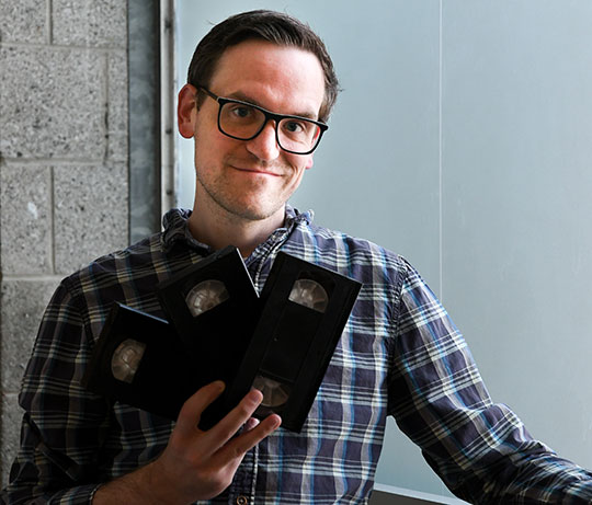 man with glasses holding VHS tapes and grinning