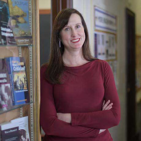 Alfred University English Professor Juliana Gray standing in a hallway with folded arms, smiling beside a bookshelf displaying literature titles.