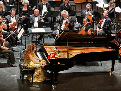 A pianist in a gold dress performs on a grand piano during a concert, with an orchestra seated behind her on stage.