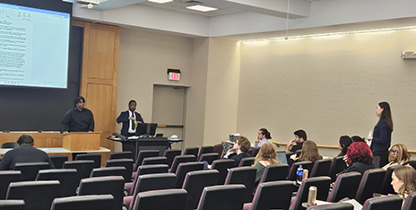 Lecture hall scene with a speaker at a podium and a presenter at a computer. Audience members sit in rows, with one standing to ask a question.