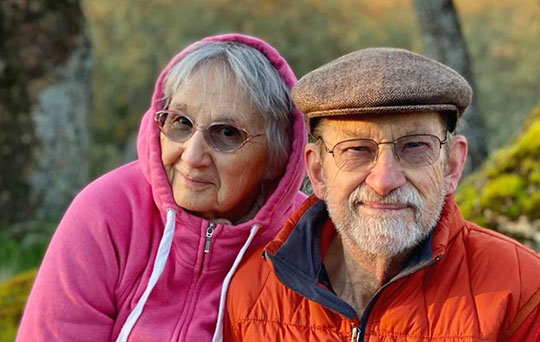 A married couple smiling warmly outdoors. The woman wears a pink hoodie, and the man in glasses has a flat cap and orange jacket. Peaceful, content expression.