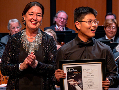 A smiling woman and a young man stand on stage during an awards ceremony; the man holds a framed certificate as an orchestra sits behind them.