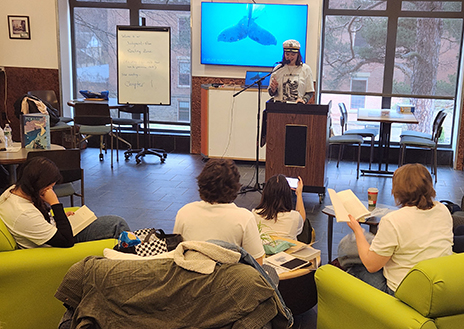 A person speaks at a podium in a classroom with a whale image on a screen behind them. Four seated people listen, reading books. Cozy and focused atmosphere.