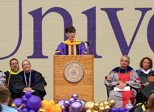 A speaker in a purple academic gown stands at a podium during a formal ceremony. Seated behind are faculty in robes. Balloons decorate the stage front.
