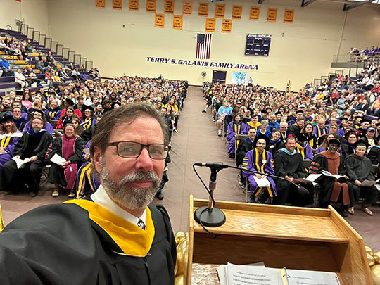 A professor in academic regalia takes a selfie at a podium in a crowded gym. Graduates in caps and gowns and faculty surround him, creating a celebratory atmosphere.