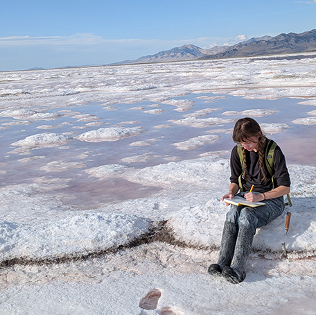 A person sits on a salt flat under a clear sky, writing in a notebook. The vast, white expanse surrounds them, evoking solitude and contemplation.