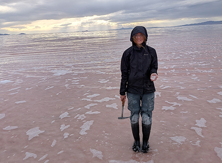 Person in a black raincoat and boots stands on a wet pinkish shoreline, holding a rock hammer, with a cloudy sky and sunset in the background. Calm and exploratory mood.