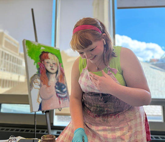 Student smiling while painting in a bright art studio with a colorful portrait on an easel behind her.