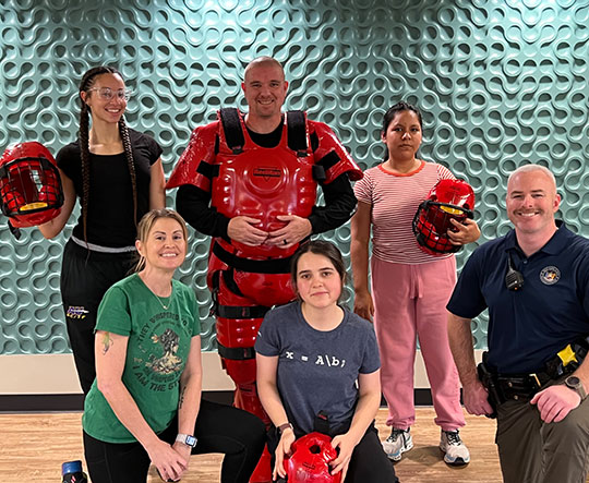 A group photo of six people in a gym, three holding red helmets. One wears red protective gear, suggesting a self-defense class. The atmosphere is positive and focused.