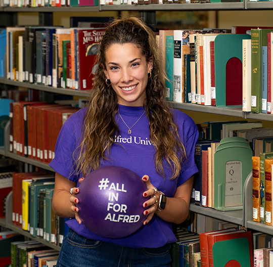 A woman in a purple "Alfred University" shirt holds a purple ball with "#All In For Alfred." She stands in a library, smiling, surrounded by colorful books.