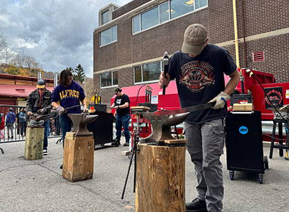 Students participate in a blacksmithing demonstration outside an Alfred University building, hammering heated metal on anvils during a campus event.