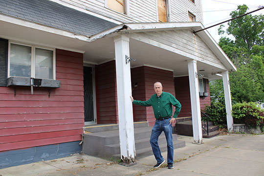 A man in a green shirt leans on a dilapidated porch column of a weathered, red multi-story building. The mood is serious, highlighting disrepair.