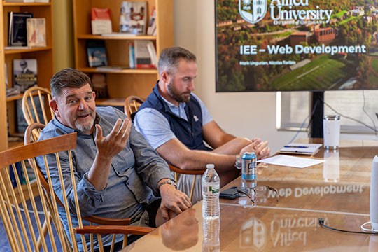 Two faculty members sit at a conference table during an Alfred University presentation on web development, with a screen displaying “IEE – Web Development” in the background.