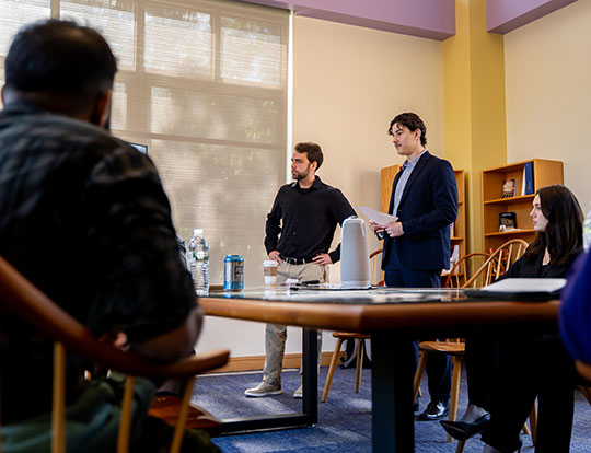 Two students stand and present during a classroom discussion while others listen around a conference table.