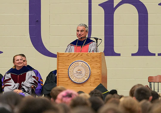 A speaker in academic regalia stands at a podium with a university seal, addressing a crowd. A seated figure nearby smiles, conveying a celebratory mood.