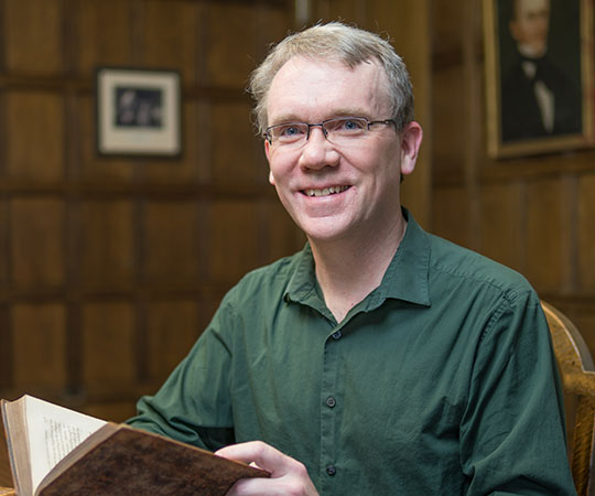 headshot of man holding a book, wearing glasses, smiling