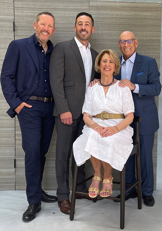 four people posing for a photo: three men standing, woman sitting on a chair
