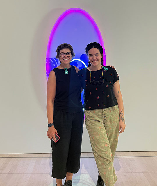 two women standing in front of a piece of neon glass art